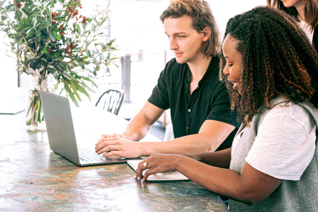 Visão de lado de um homem branco e uma mulher negra trabalhando em uma mesa de madeira. Ele está mexendo em um computador e ela está fazendo anotações em um caderno.