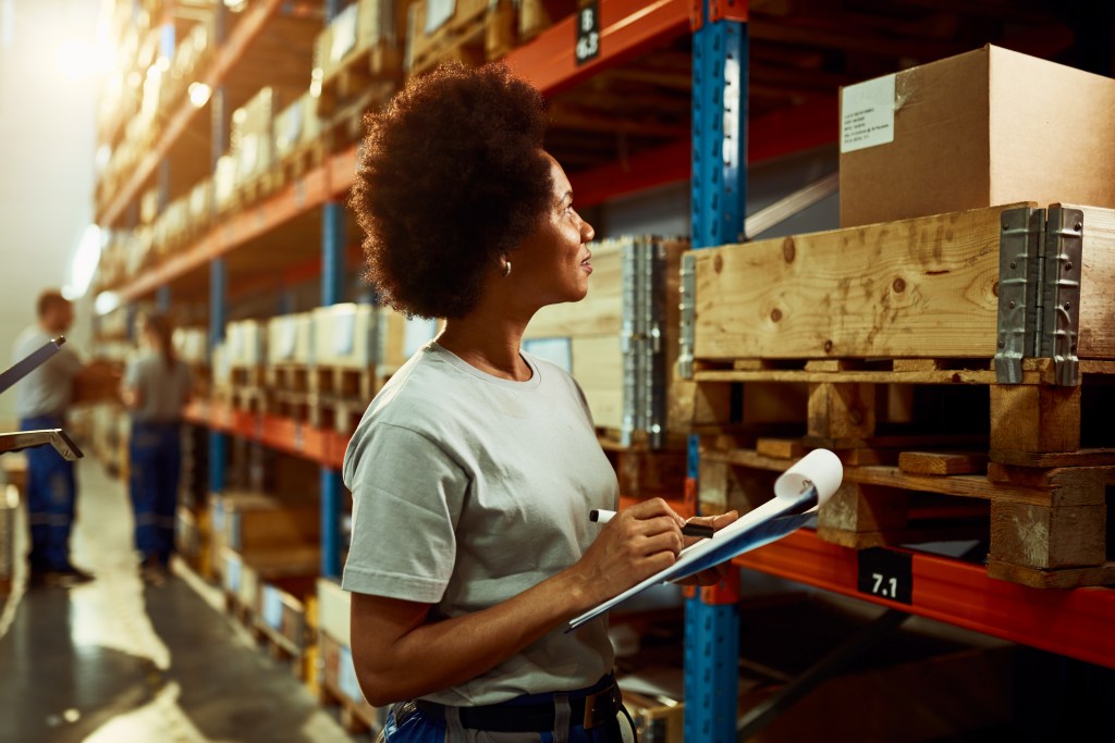 Mulher afroamericana com uma camiseta branca, com uma prancheta e uma caneta na mão, olhando para uma prateleira cheia de caixas em um centro de distribuição.
