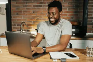 Um homem negro está sorrindo, sentado em uma mesa de madeira mexendo no notebook.